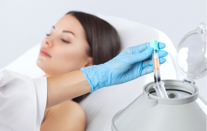 Taking a tube out of a centrifuge with the plasma and platelets now separated from the blood sample to create PRP to be used on the female patient laying flat in the background