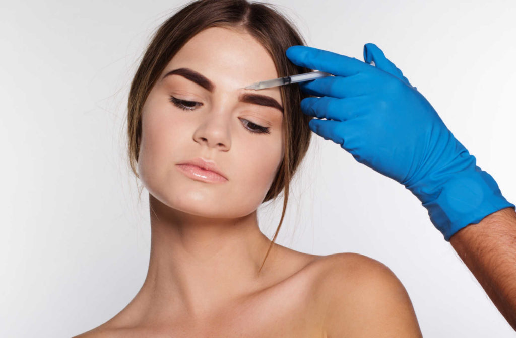 A hand wearing a blue medical glove injecting botox on a woman's face for a botox brow lift.