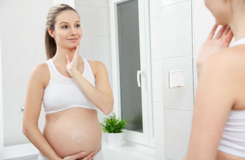 A pregnant women looking in the mirror while touching her cheek, noticing how her skin is changing during pregnancy.