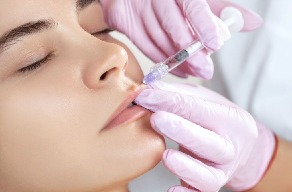 A woman receiving dermal fillers in her lips from an aesthetician wearing pink medical gloves.