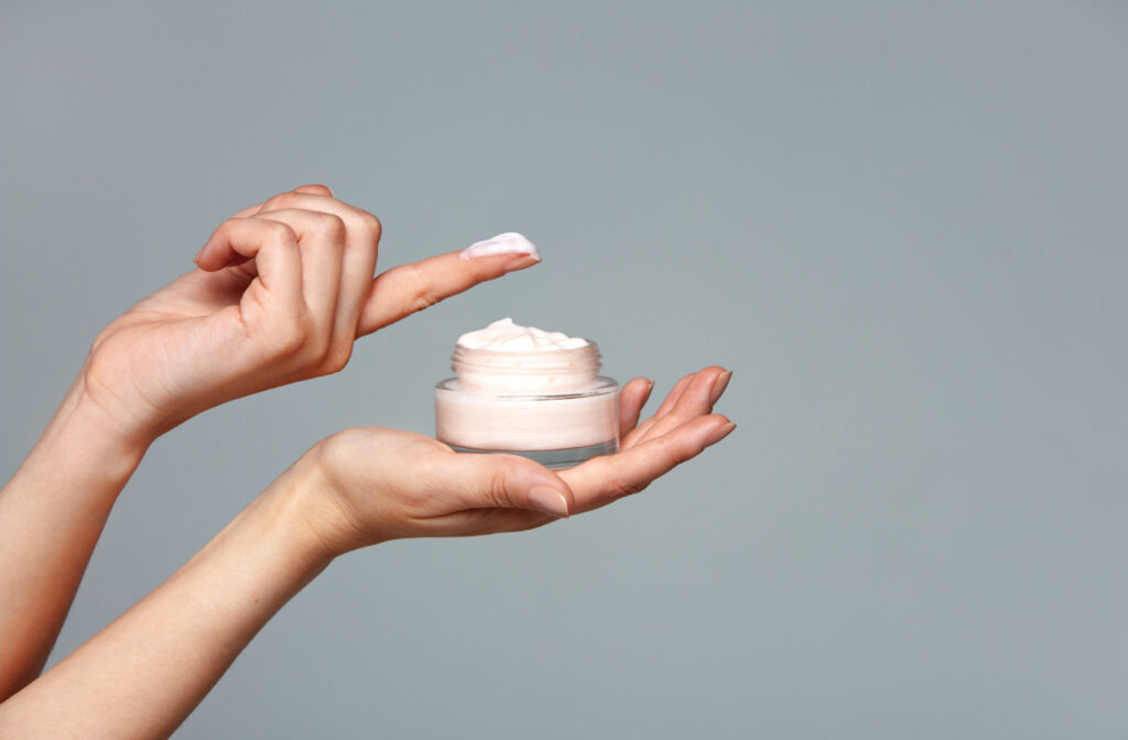 A hand holding a jar of cream and the other hand has a small amount of cream on the pointer finger.