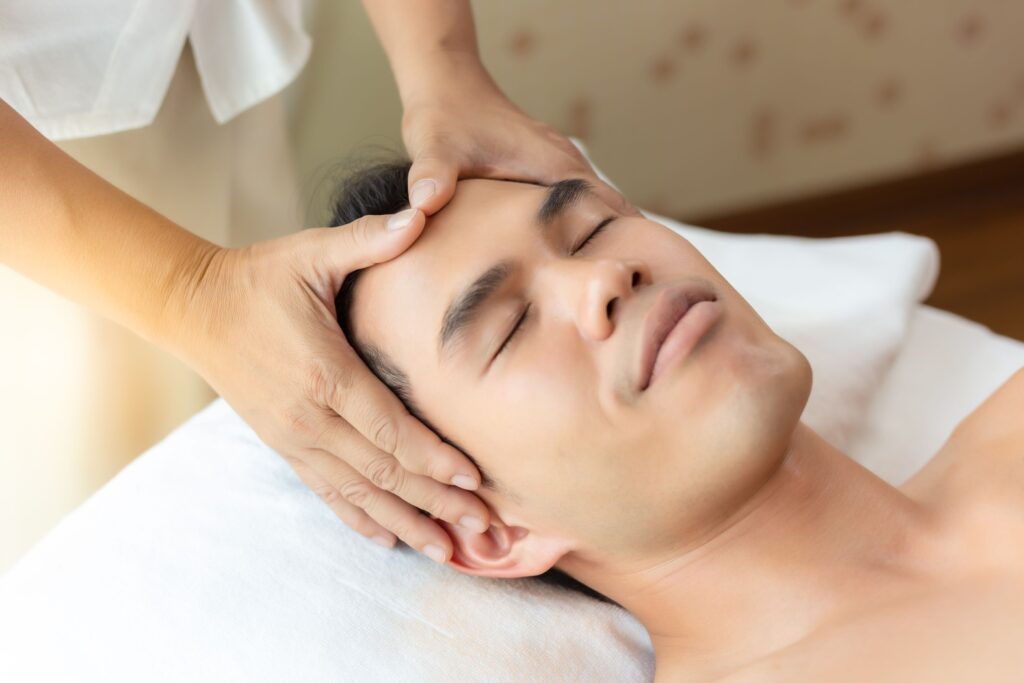 A young man is laying down with his eyes closed while he is receiving a facial.