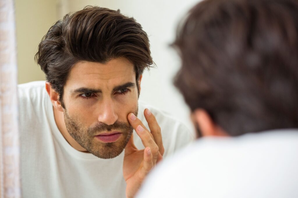 A young man focusing on his reflection in the mirror while touching his face.