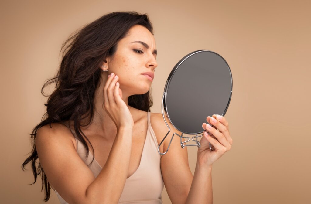 An aesthetics patient in front of a brown background using a handheld mirror to examine their skin.