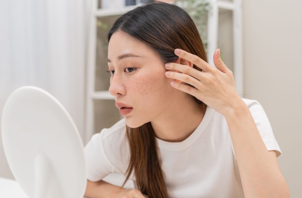 A young adult checking their skin in a small mirror to look for signs of sun damage.