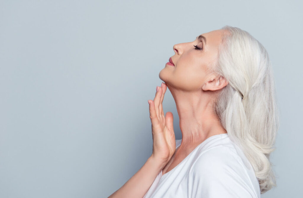 An older patient in front of a gray background tilting their chin high and touching their wrinkle-free neck.