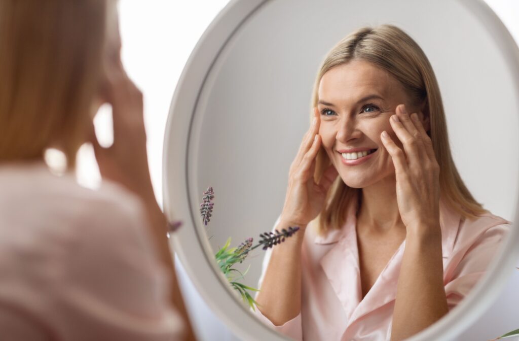 A young adult smiling in a mirror and touching their face after skin tightening treatment.